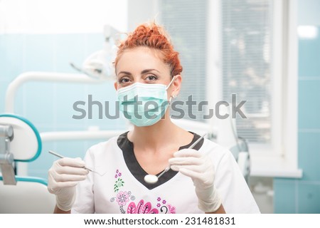 Half-length portrait of young dentist wearing medical mask and gloves standing holding tweezers and little mirror wanted to examine her patient