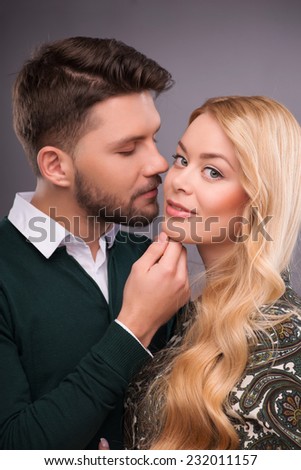 Half-length portrait of handsome young man wearing white shirt and dark-green cardigan standing hugging his beautiful fair-haired girlfriend wanted to kiss her