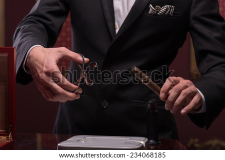 Selective focus on the hands of young aristocrat wearing costly black official costume wanted to cut the tip of a Cuban cigar