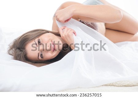 Half-length portrait of dark-haired beautiful smiling girl wearing sexy lingerie lying in the bed playing with the bedclothes thinking about her boyfriend