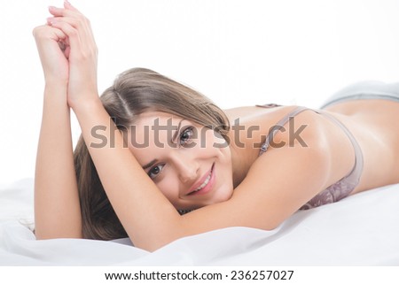 Selective focus on the dark-haired beautiful smiling girl wearing sexy lingerie lying in the bed on her belly looking at us dreaming about her future date