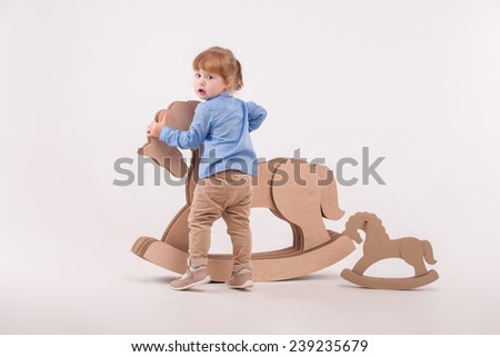 Full-length portrait of little lovely girl wearing blue shirt and brown pants standing back to us holding at the wooden toy horse. Isolated on the white background