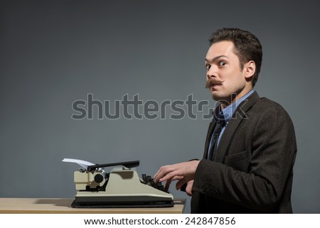 Just inspired . Young nerd author typing something at the typewriter and looking at camera while sitting at his working place against grey background