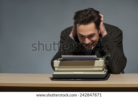 Author creating. Handsome young author working at the typewriter and ruffling his hair while sitting at his working place against grey background