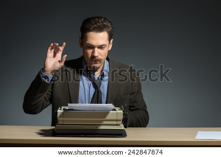 Inspired author. Handsome young author working at the typewriter raising hand while sitting at his working place against grey background