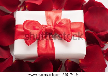 Gift to beloved. Top view closeup image of white present box with red ribbon on top decorated with rose petals on white background
