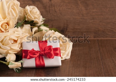Gift to the dearest. Closeup image of beautiful bouquet of white roses arranged with a white gift box tied with red ribbon and placed on wooden background