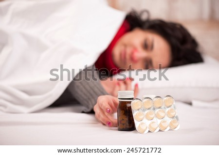 She is unwell. Young sick woman lying in bed with red nose in thick scarf and trying to take medicines and lozenges