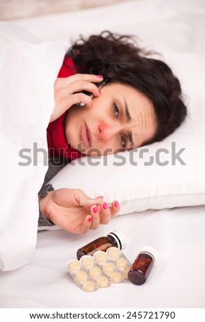 On sick leave. Young sick woman in thick scarf talking over the telephone and holding thermometer while lying in bed and having medicines and lozenges on the blanket