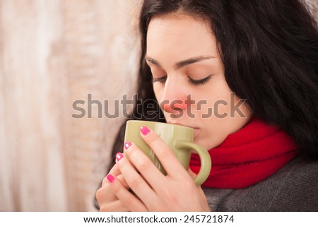 Sick woman with cup of tea. Closeup image of young frustrated woman in knitted scarf suffering from flu and holding a cup of tea while sitting against wooden wall
