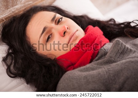 Sick woman with virus. Closeup image of depressed young woman with red nose lying in bed with thick scarf on her neck and looking upset