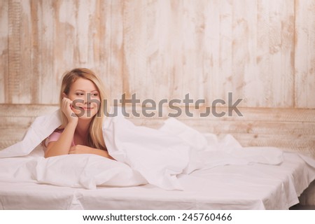 Day dreaming in bed. Attractive young smiling woman lying in bed and touching her face with hand while looking away