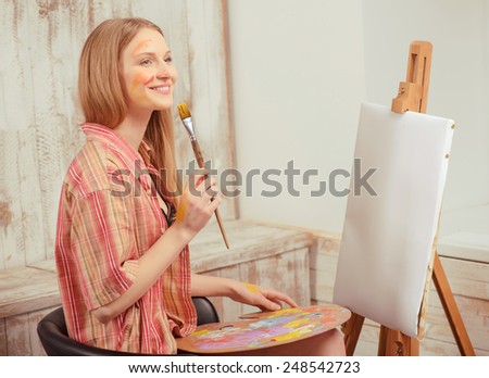 Beautiful painter. Young beautiful woman in a checked shirt and face colored with paint in front of the blank drawing easel sitting against artistic working place