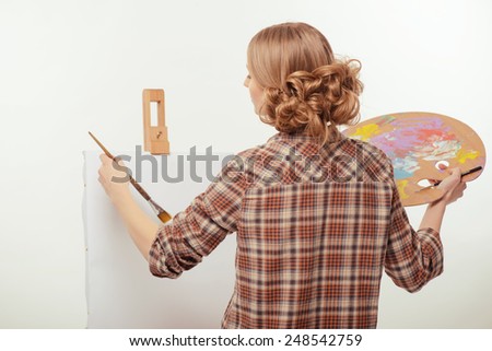 Creative hobby. Rear view of young beautiful woman painting at the drawing easel with a paintbrush at copy space and holding a palette while standing isolated against white background