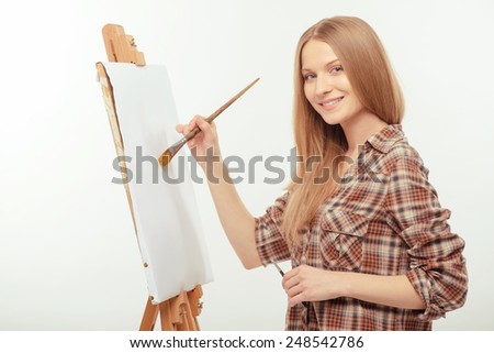 The inspiration is somewhere around. Young beautiful woman in a checked shirt painting with a brush on drawing easel with copy space while standing isolated against white background