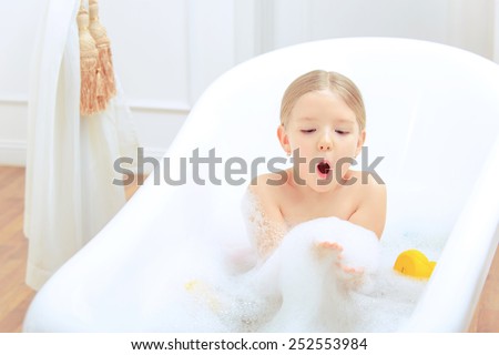 Bath time is fun. Top view image of a cute little girl taking a bath and blowing soap foam while sitting on a luxurious bathtub with rubber ducks