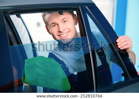 Welcome to our car service station. Closeup image of a handsome car mechanic wiping the car windows with tinting foil and smiling at camera in specialized service station