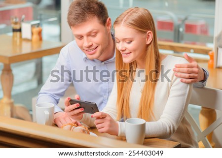 No minute without technologies. Beautiful young loving couple bonding to each other and looking at mobile phone while sitting in cafe together