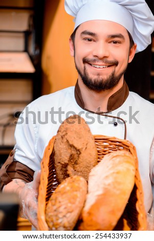 Proud of his baked goods. Cropped image of a handsome young man in apron holding basket with baked goods in bakery shop