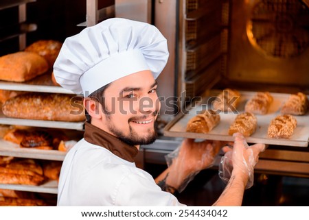 Fresh baked bread. Confident young man in apron taking the fresh baked bread from oven and looking over his shoulder with a smile in the bakery shop