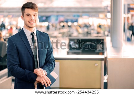 Timely check-in. Handsome young businessman in suit holding his passport with ticket and luggage while standing against airline check in counter in the airport