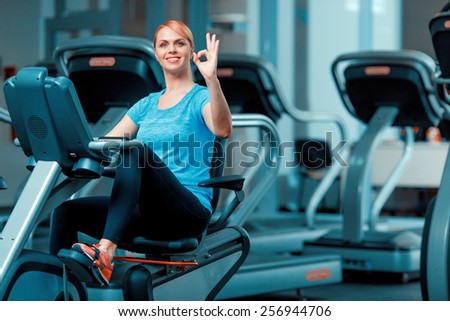 Cycling on exercise bike. Beautiful mature woman in sports clothing working out on exercise bicycle in the gym and showing ok sign