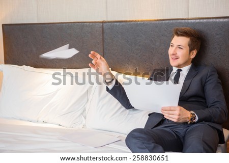 Enjoying business trips. Confident young businessman in suit and tie reading documents and playing with paper plane while sitting on the bed in luxury hotel room