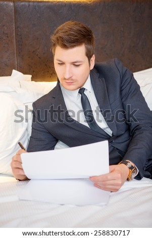 He likes business trips. Confident young businessman in suit and tie reading documents while sitting on the bed in luxury hotel room