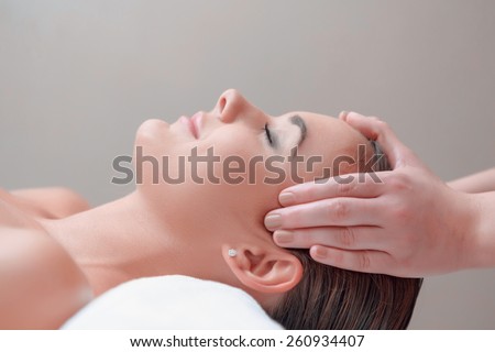 Facial massage. Top view of attractive young woman lying on back and keeping her eyes closed while massage therapist massaging her face