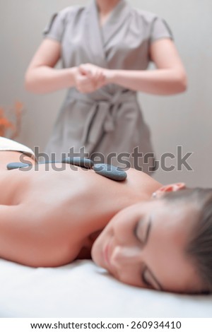 Stone SPA. Top view of young woman with shiny skin lying on front with spa stones on her back and smiling massage therapist on the background