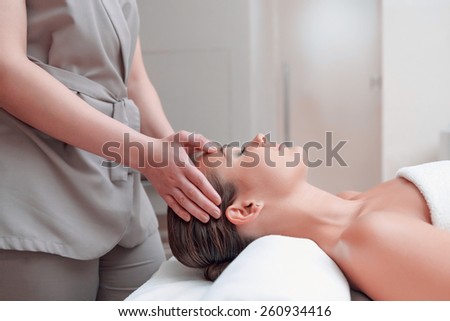 Facial massage. Top view of attractive young woman lying on back and keeping her eyes closed while massage therapist massaging her face