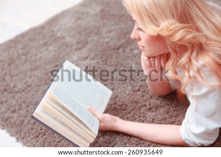 She found a peaceful place to read. Top view of confident female student reading a book while lying on the carpet