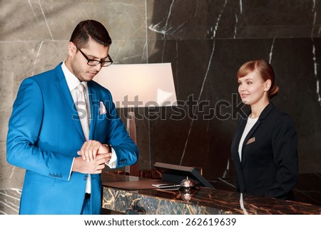 Time is money. Businessman in blue suit looks at his watch standing just in front of the reception desk with smiling receptionist behind