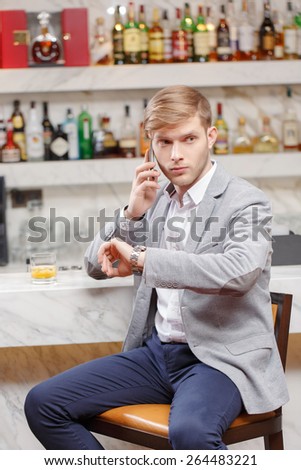 Anticipating a meeting. Young man in shirt and jacket talking over the phone and looking at his watch while sitting at the counter of the bar