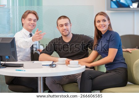 Fancy this beauty. Young smiling salesman points with the hand to attract attention of the couple willing to buy a car. Computer, keys and clipboard with documents lay on the table