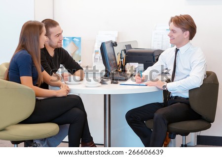 Sorting out car finances. Young smiling sales consultant communicates with a couple sitting by the table with computer, printer and office stationery on it and tells technical information of a car