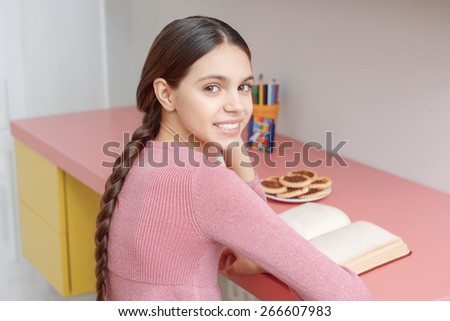 Exciting reading. Cute teenage girl with long dark hair reading a book at the table turns and smiles to the camera