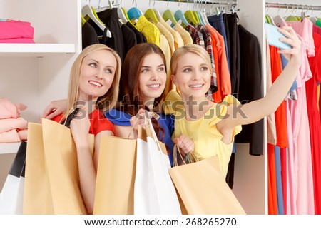 Black Friday. Three female shoppers smiling happily to the camera of their phone holding numerous paper bags in a fashion store