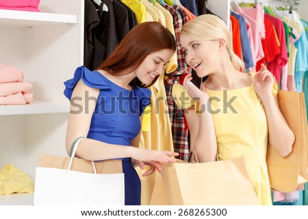 Really cool clothes here. Young smiling shopper speaking over the phone while her friend is peeping inside her paper bags copyspace