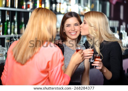 Friends love each other. Touching shot of a blond woman kissing on the chick her female friend who joyfully laughs holding a cocktail in the bar