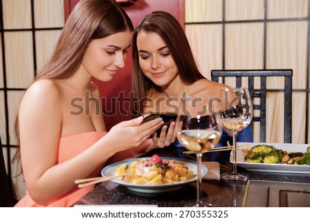 Sharing photos with friend. Two beautiful women wearing dresses have a dinner at restaurant and looking on the screen of the smartphone
