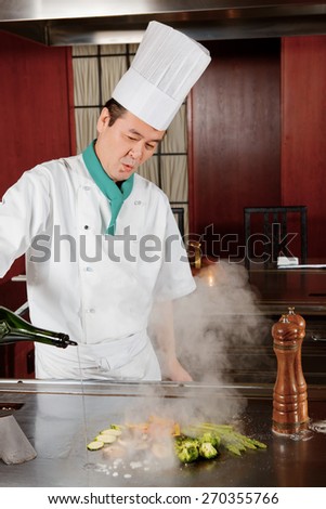 Spice cuisine. Japanese cook in uniform pouring sauce on fried vegetables on hot oven