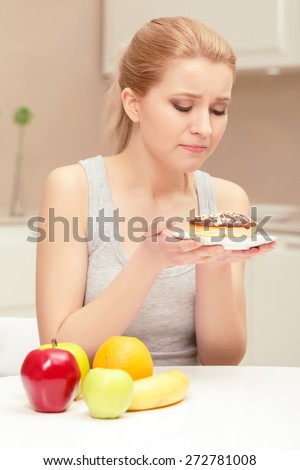 Feeling a temptation. Nice young woman feeling desire to eat a donut and looking on it but banana, oranges and apples lying on the table remind her of eating healthy food