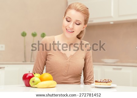 Smiling and healthy. Young happy and glad woman sitting at the table looking on the plate with fruit prefers apples, oranges and banana to donut