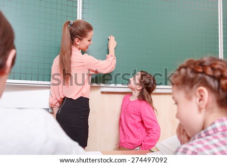 I will write. Smiling female teacher and pupil standing at blackboard, teacher is going to write on blackboard.