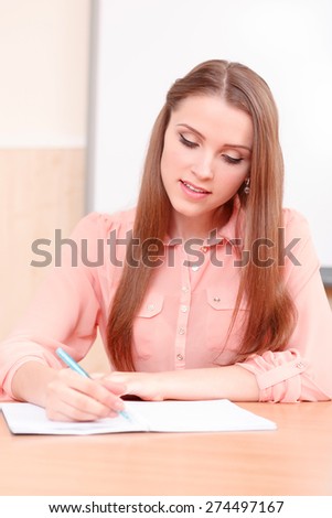 Diligent student. Youthful attractive woman writing in copy book on background of white blackboard.