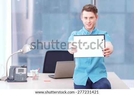 Look at this. Young smiling businessman sitting on desk and holding folder with papers and pencil.