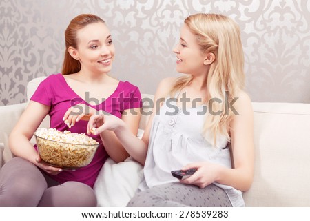 Movie night. Two young beautiful girls sitting on a white couch discussing a movie while holding remote control and eating popcorn at pajama party