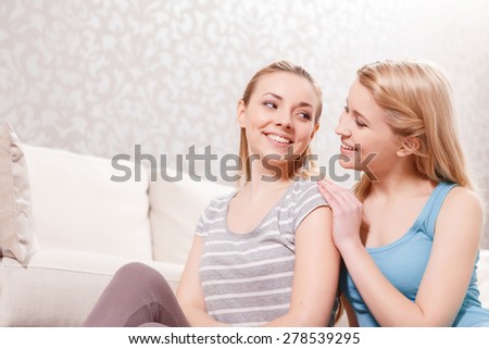 Friendship. Two young beautiful blond girls wearing pajamas sitting on the floor near the couch and looking at each other smiling