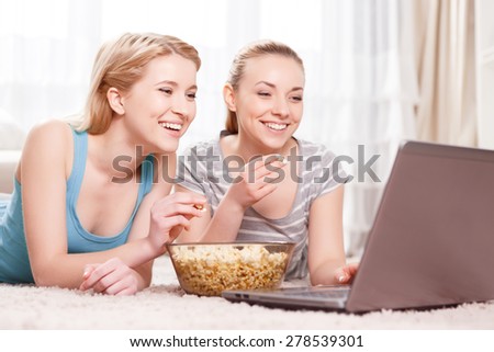 Pajama party. Two young beautiful blond girls wearing pajamas lying on the floor eating popcorn and looking at the laptop